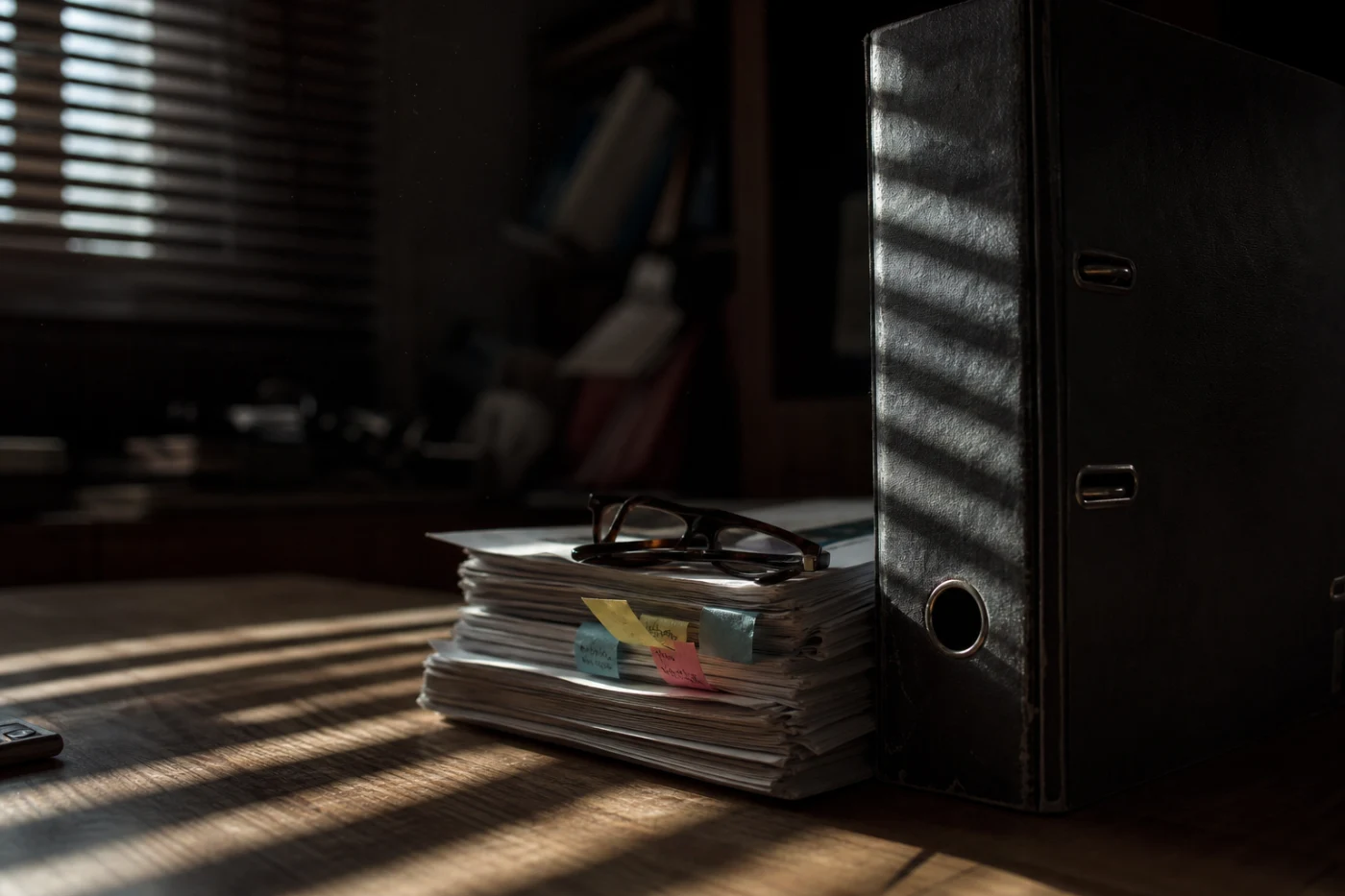A lever-arch compliance binder with uneven coloured index tabs, striped by light through a slatted blind.