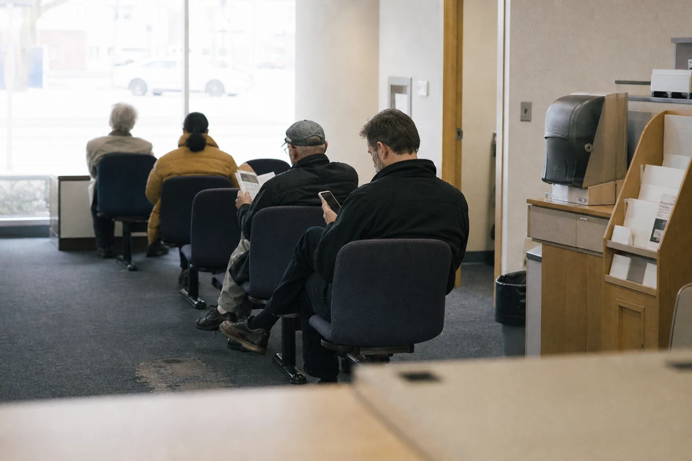 The waiting area of a small branch mid-morning, several people seated on bolted chairs, nobody interacting.