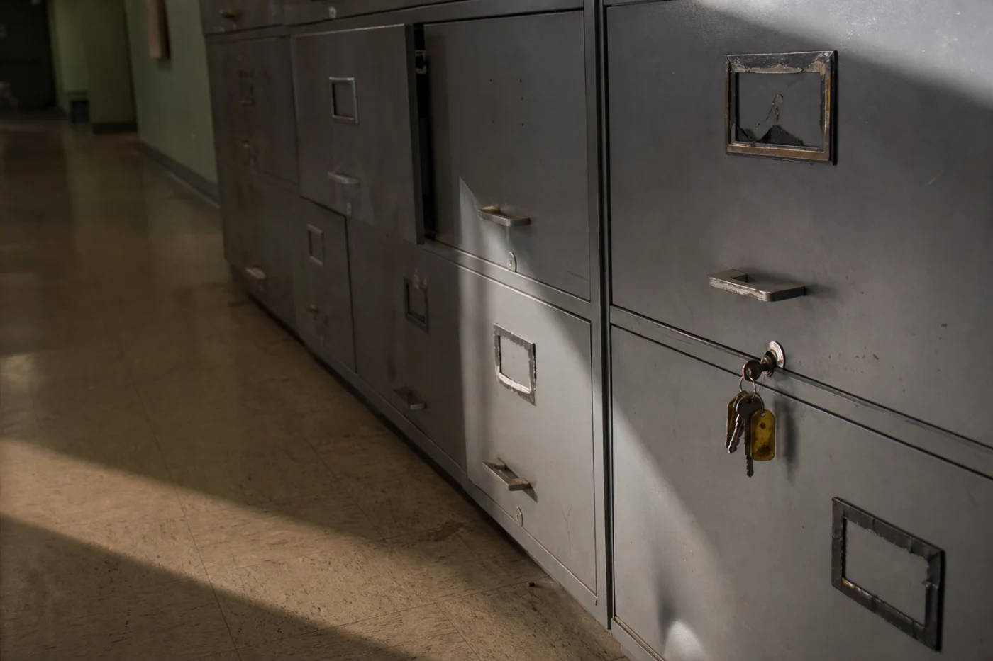 A row of locked filing cabinets along a clinic corridor, keys hanging from one lock in hard afternoon light.