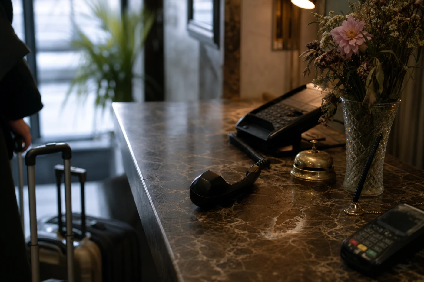 A hotel reception counter with the desk phone off its cradle, suitcase handles and a coat sleeve at the frame edge.