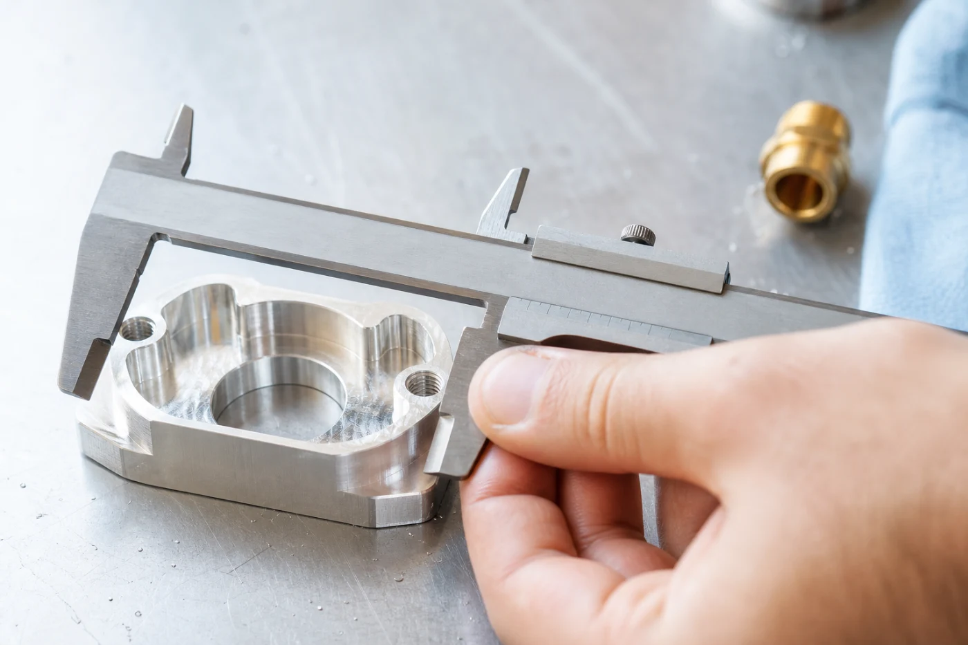 Vernier calipers closed around a machined aluminium component on a steel bench, a hand steadying the part.