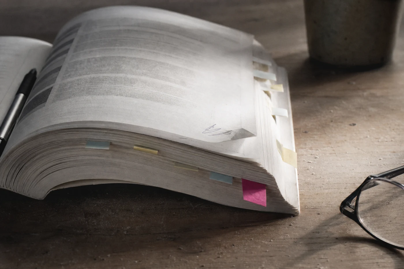 A printed monthly report lying open on a studio table, pages tabbed with coloured flags, reading glasses folded at the edge.