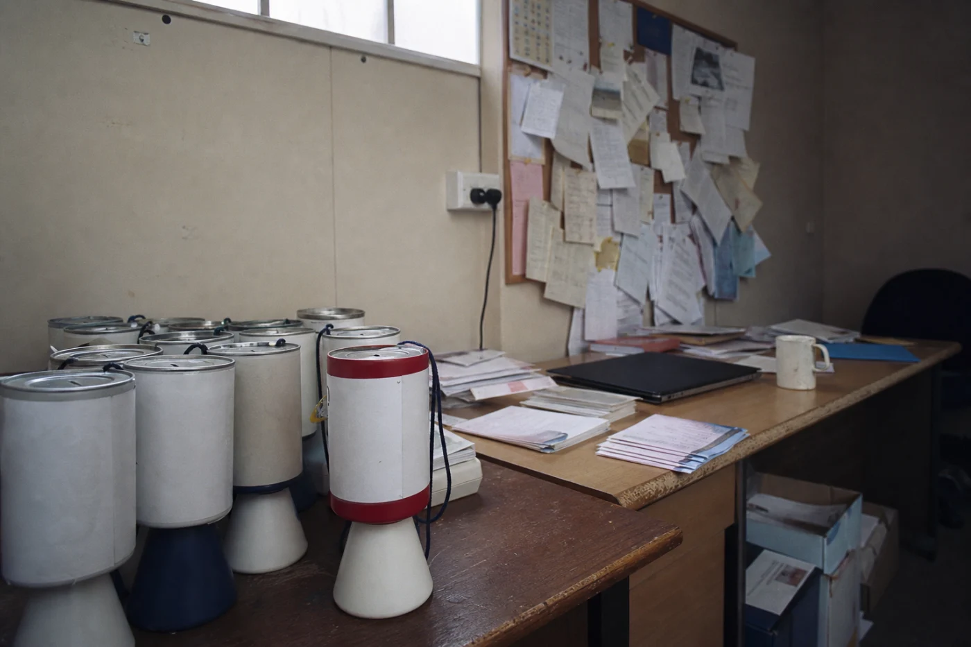 A small charity office with two mismatched desks, sealed collection tins stacked on one and folded leaflets on the other.