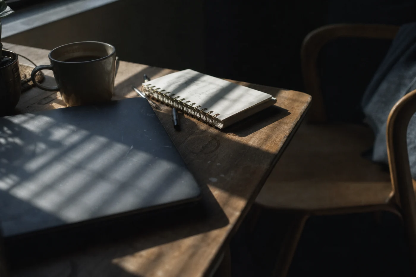A laptop closed mid-task beside a notebook with a half-written list, hard morning light striping the lid.