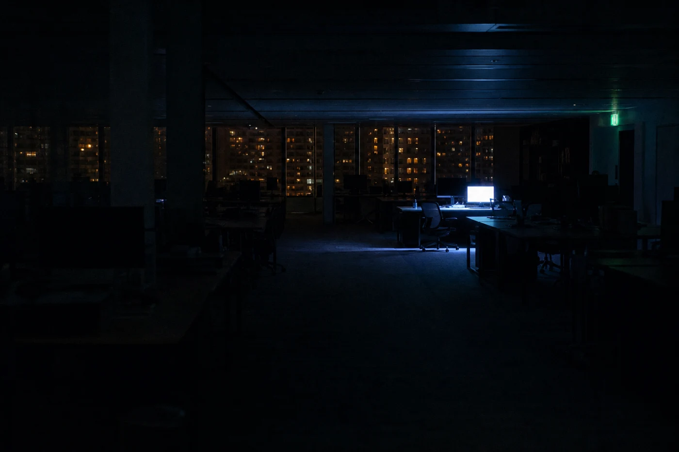 An empty open-plan office at night, one monitor left awake throwing a cold pool of light across a single desk.
