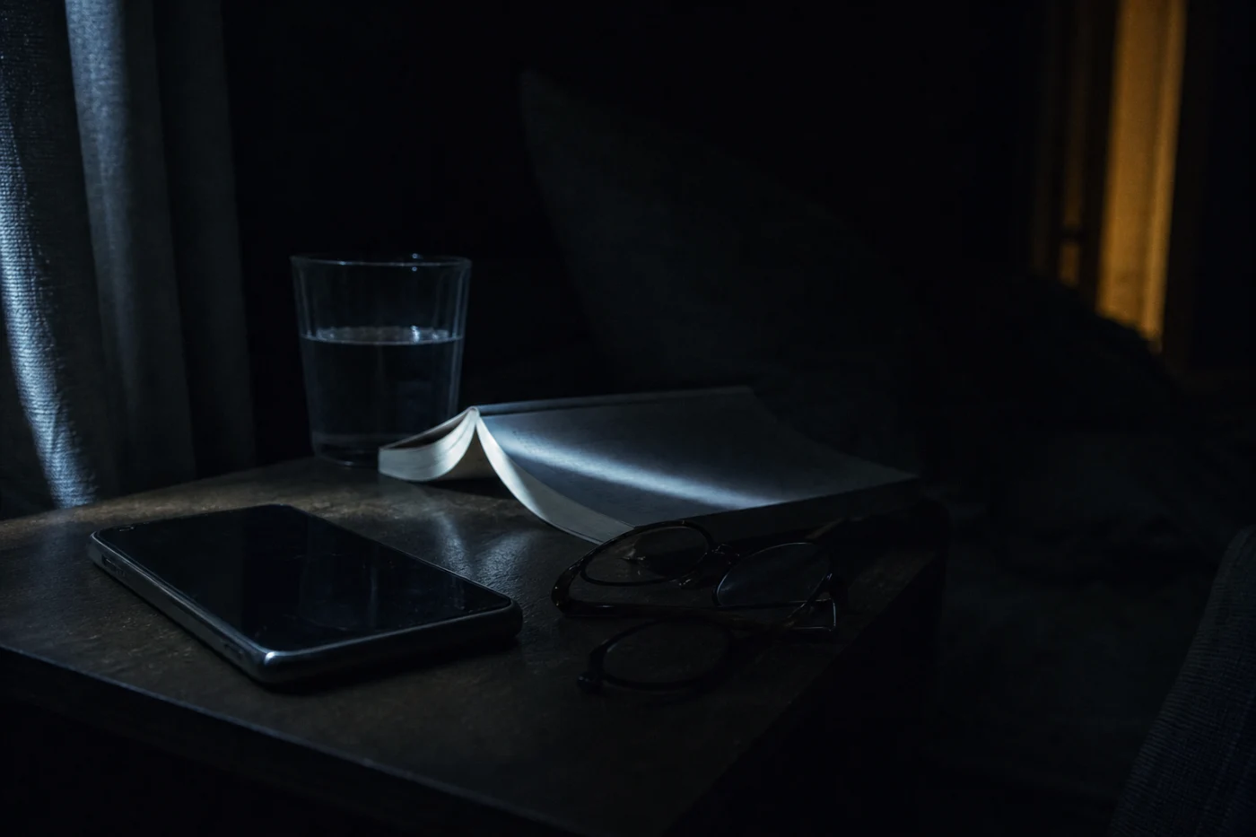A bedside table in the small hours: a dark phone, a paperback face-down, a glass of water, cut by a hard band of streetlight.