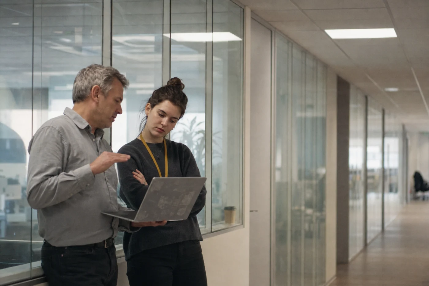 Two colleagues at a glass partition wall, one balancing a laptop on his forearm mid-explanation while a newer colleague listens.