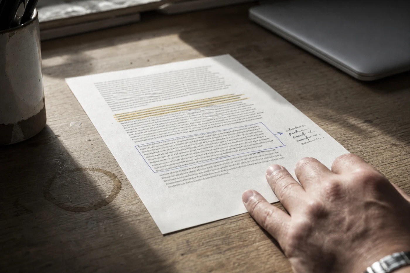 A printed article on a desk in hard window light, one paragraph run through with yellow highlighter and another boxed in pen with an arrow to a handwritten margin note.