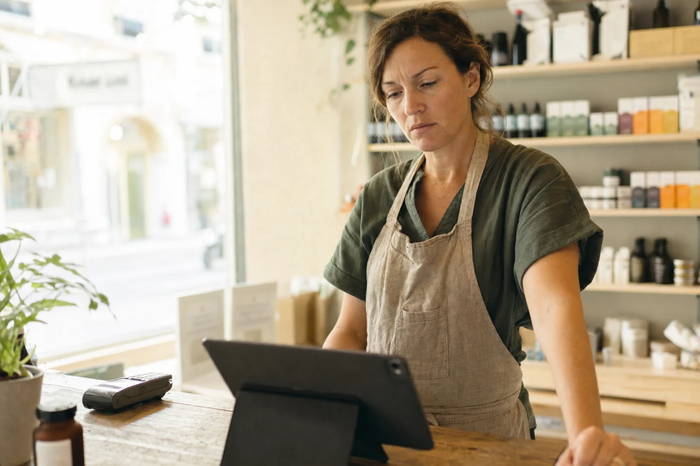 A shop owner standing behind her counter during a quiet moment, reading a tablet with a thoughtful, unconvinced expression.