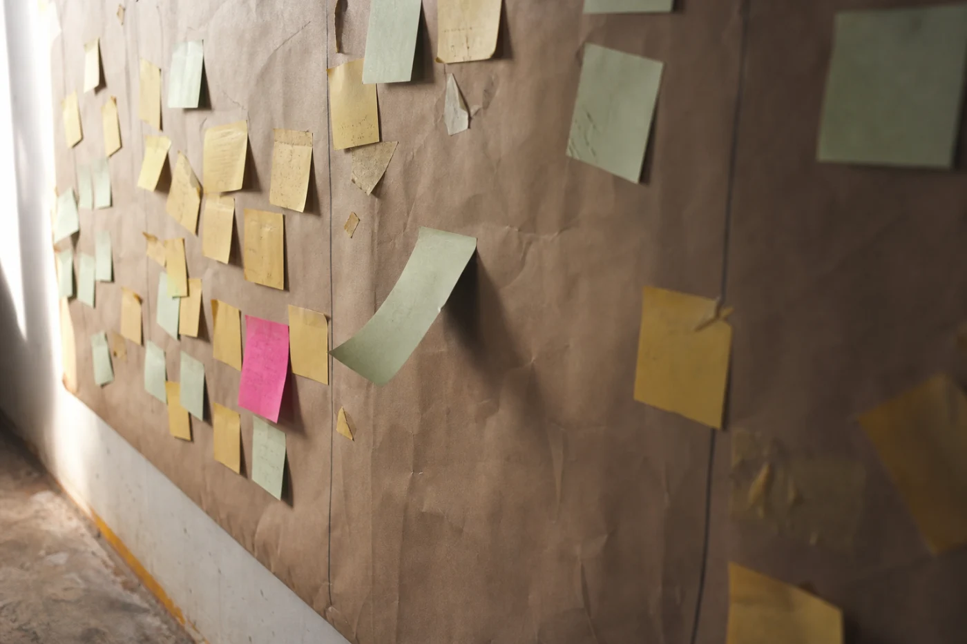 A studio wall of kraft paper covered in sticky notes in rough columns, raking side light throwing a shadow from each one.